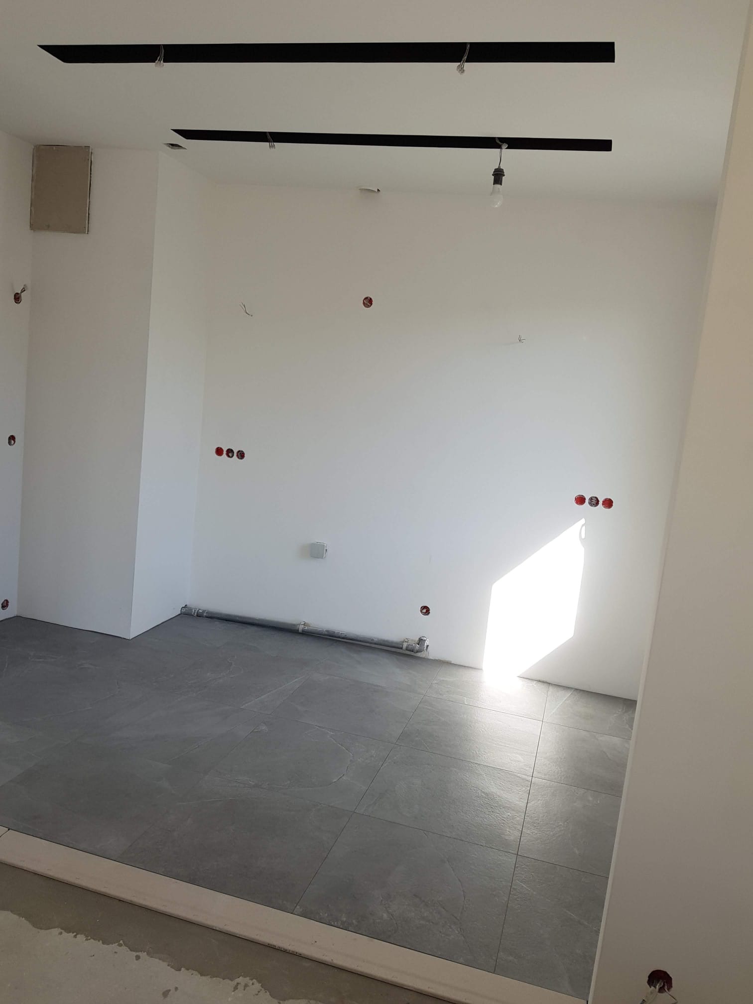 Wnętrze w trakcie prac: szare płytki podłogowe, białe ściany z widocznymi puszkami elektrycznymi i rurami. Na suficie czarne listwy oświetleniowe.