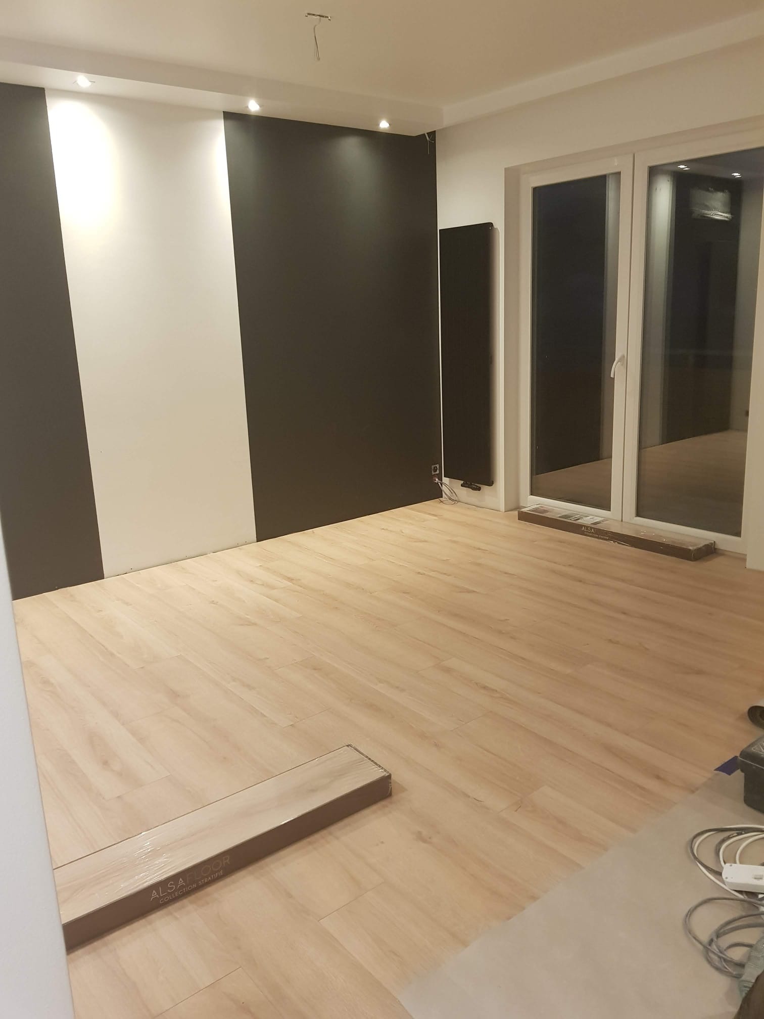 Jasne panele podłogowe w pokoju z oknem balkonowym i ścianą w biało-czarne pasy. Widoczny karton z panelami i narzędzia.