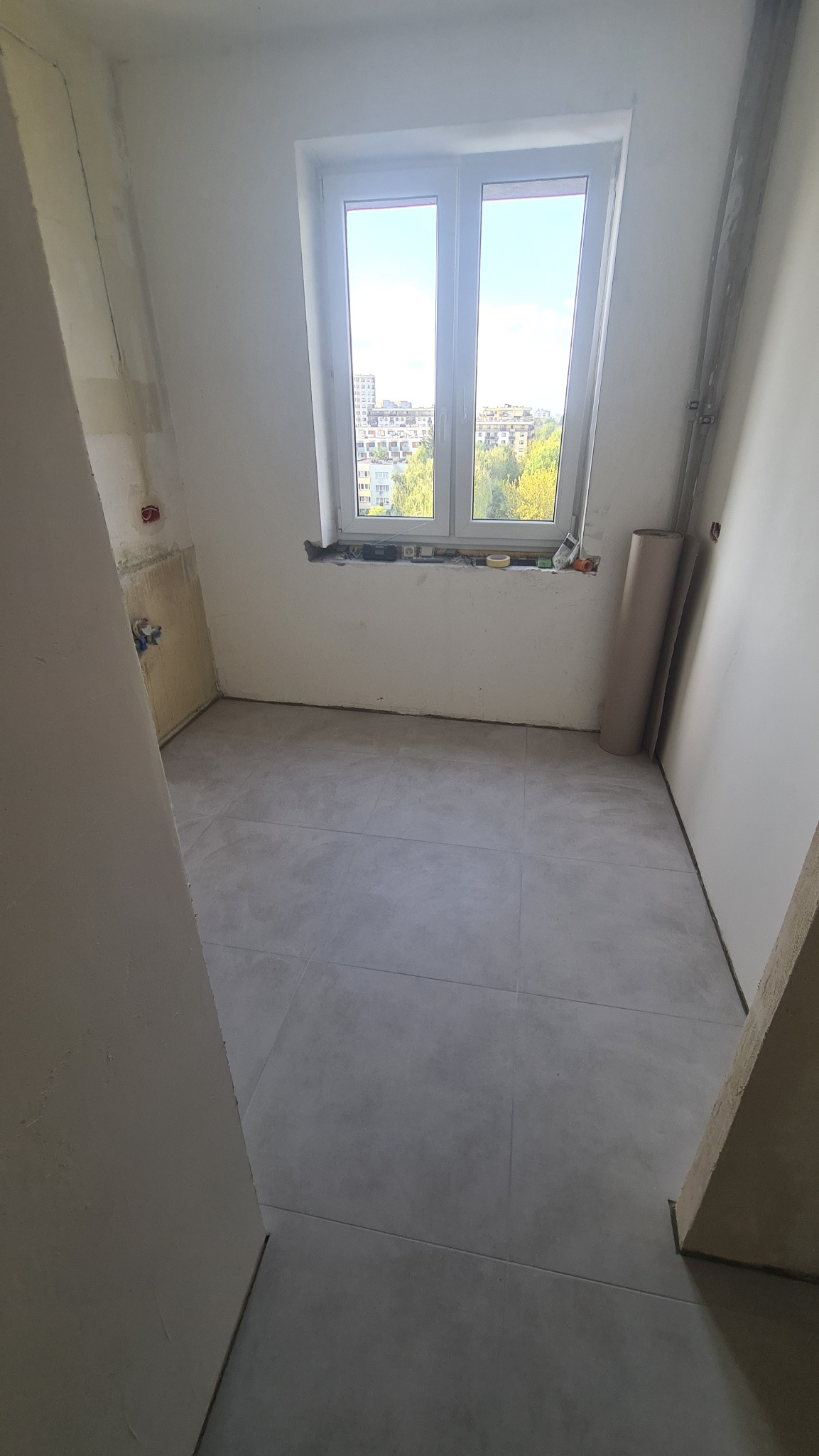 Jasne płytki podłogowe w pokoju z oknem i widokiem na miasto. Ściany przygotowane do dalszych prac wykończeniowych. Rolka papieru zabezpieczającego pod ścianą.