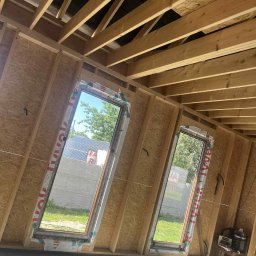 Okna24Expert - Montaż okien dachowych w surowym wnętrzu z drewnianą konstrukcją. Widok na zewnątrz przez okna, narzędzia pracy obok.