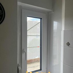 Okna24Expert - Białe okno PCV z widokiem na zewnątrz, zamontowane w jasnym pomieszczeniu z białymi płytkami. Widoczna roleta zwinięta na górze okna.