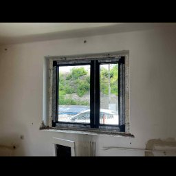 Okna24Expert - Czarne okno PCV z widokiem na zieleń, osadzone w surowym otworze okiennym. Widoczne ślady po montażu i niedokończone wykończenie ściany.