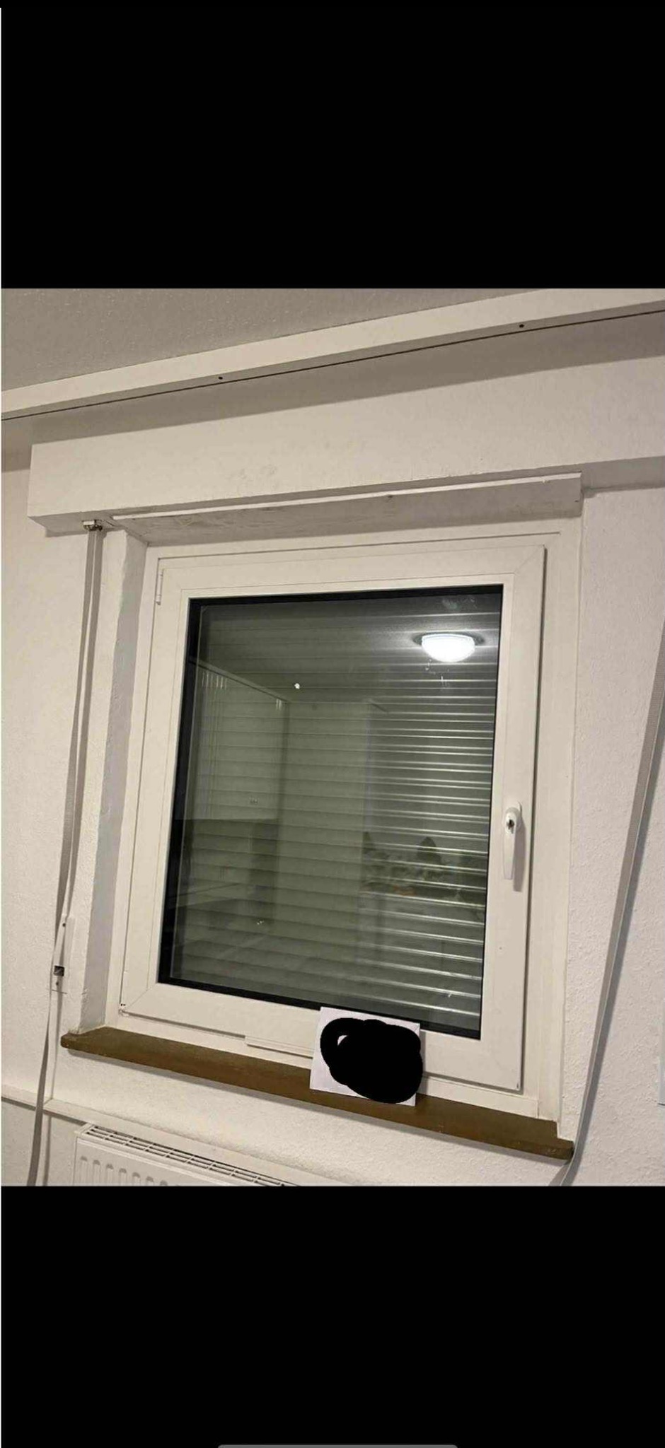 Białe okno PCV z żaluzjami wewnętrznymi, widoczne z bliska. Na parapecie kartka z zamazaną treścią. Nad oknem zabudowana roleta.