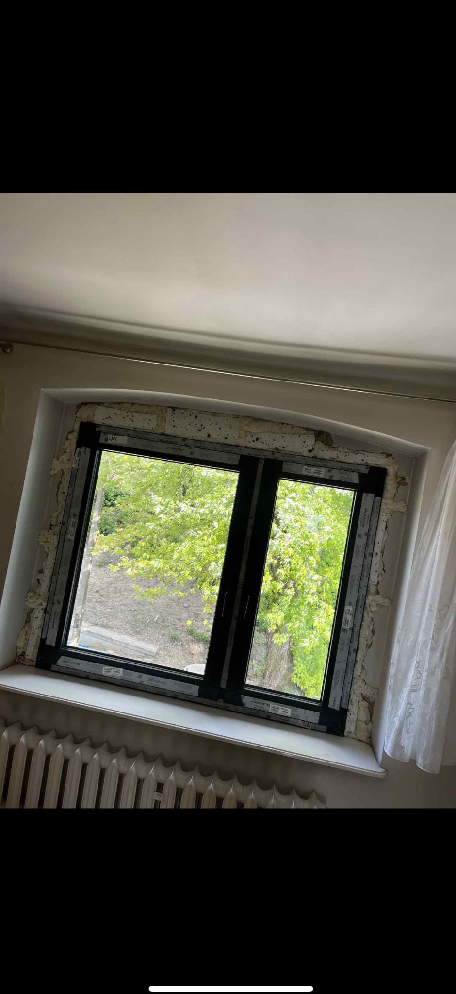 Czarne okno PCV Salamander po montażu, uszczelnione pianką montażową, z widokiem na zieleń drzew za oknem. Grzejnik pod oknem.