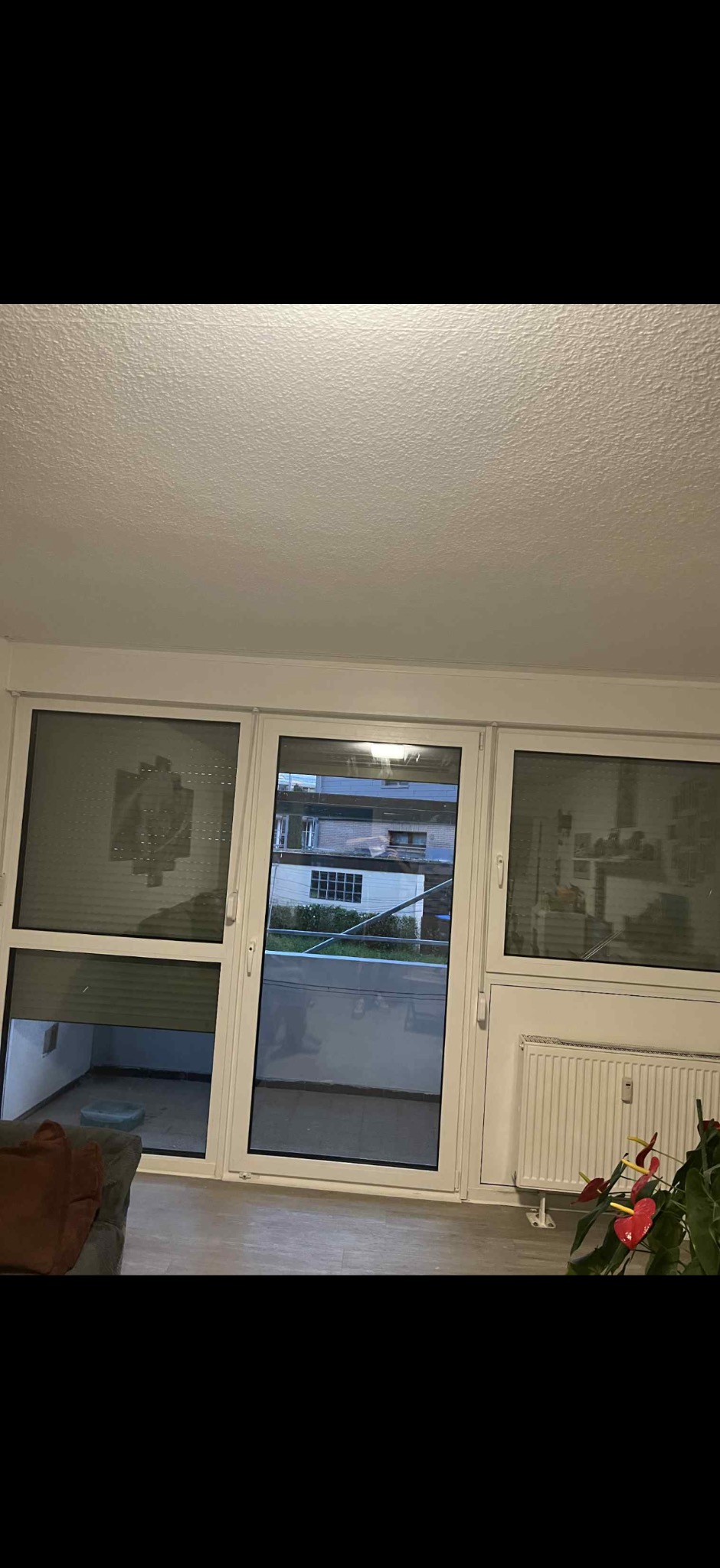 Białe okno balkonowe z roletą w połowie opuszczoną, widok na balkon i budynki na zewnątrz. Wnętrze z grzejnikiem i kwiatem doniczkowym.