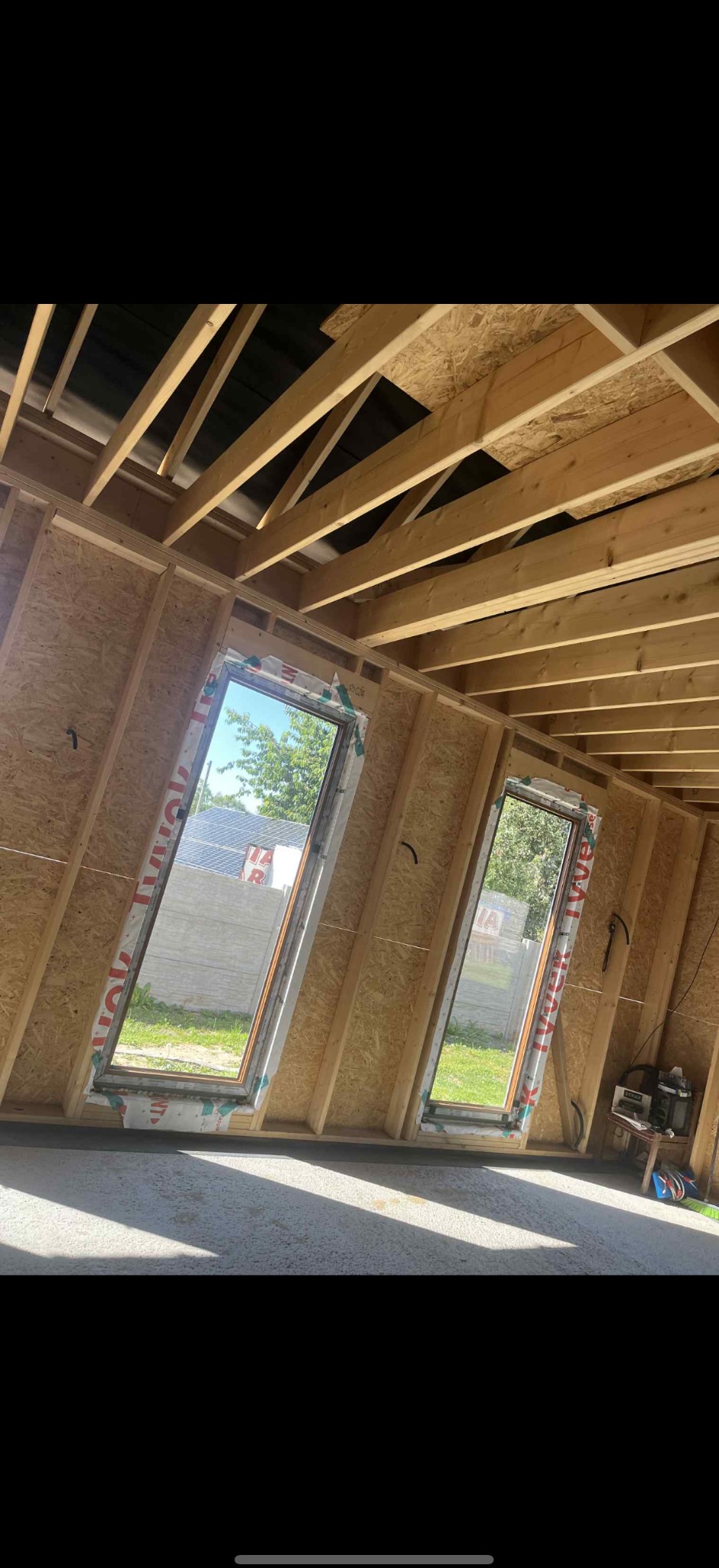 Montaż okien dachowych w surowym wnętrzu z drewnianą konstrukcją. Widok na zewnątrz przez okna, narzędzia pracy obok.