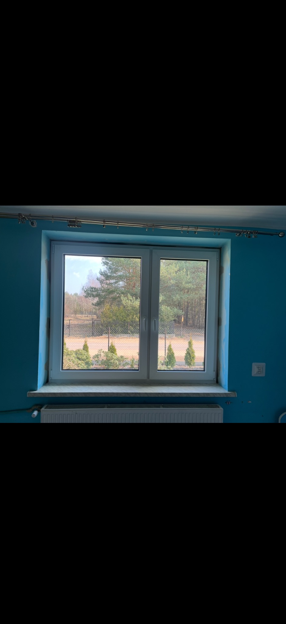 Białe, dwuskrzydłowe okno PCV z widokiem na zewnątrz, gdzie widać drzewa i krzewy. Ściana w pokoju pomalowana jest na niebiesko. Pod oknem zamontowany jest grzejnik.