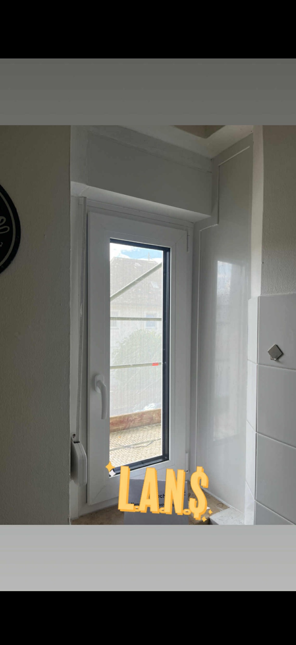 Białe okno PCV z widokiem na zewnątrz, zamontowane w jasnym pomieszczeniu z białymi płytkami. Widoczna roleta zwinięta na górze okna.