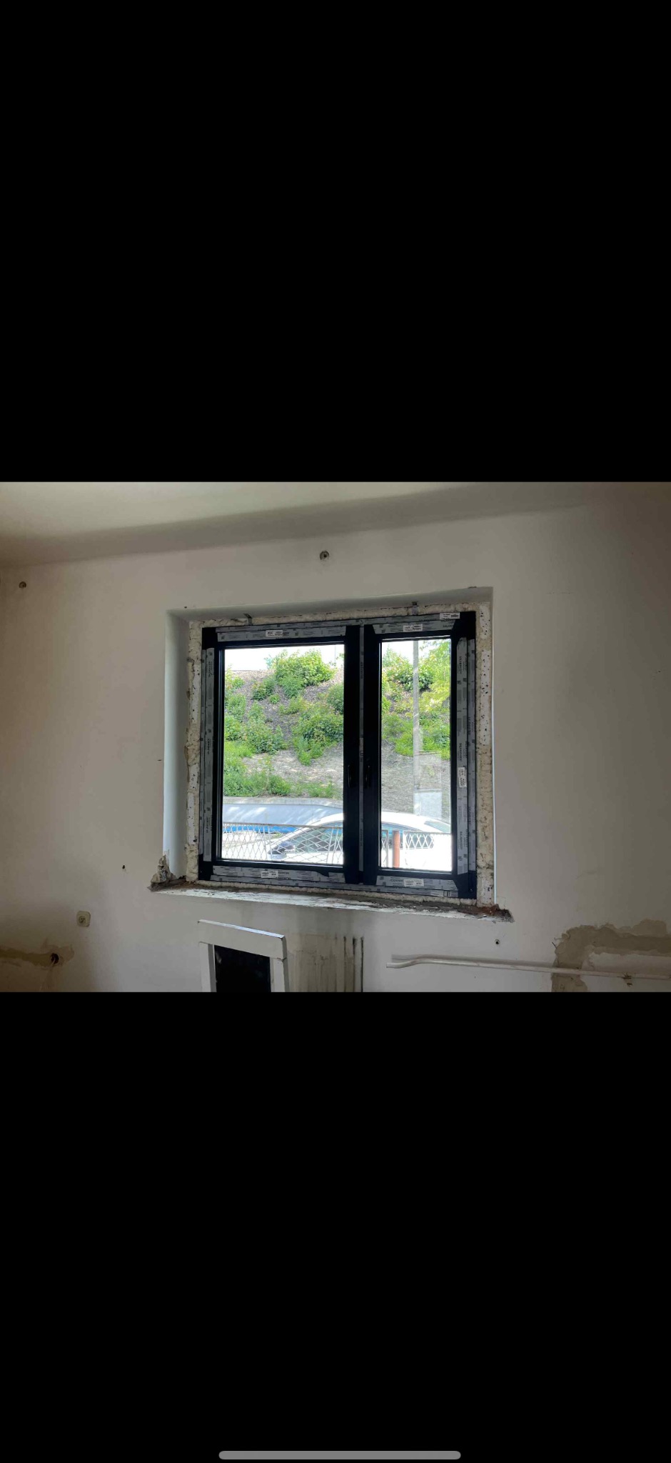 Czarne okno PCV z widokiem na zieleń, osadzone w surowym otworze okiennym. Widoczne ślady po montażu i niedokończone wykończenie ściany.