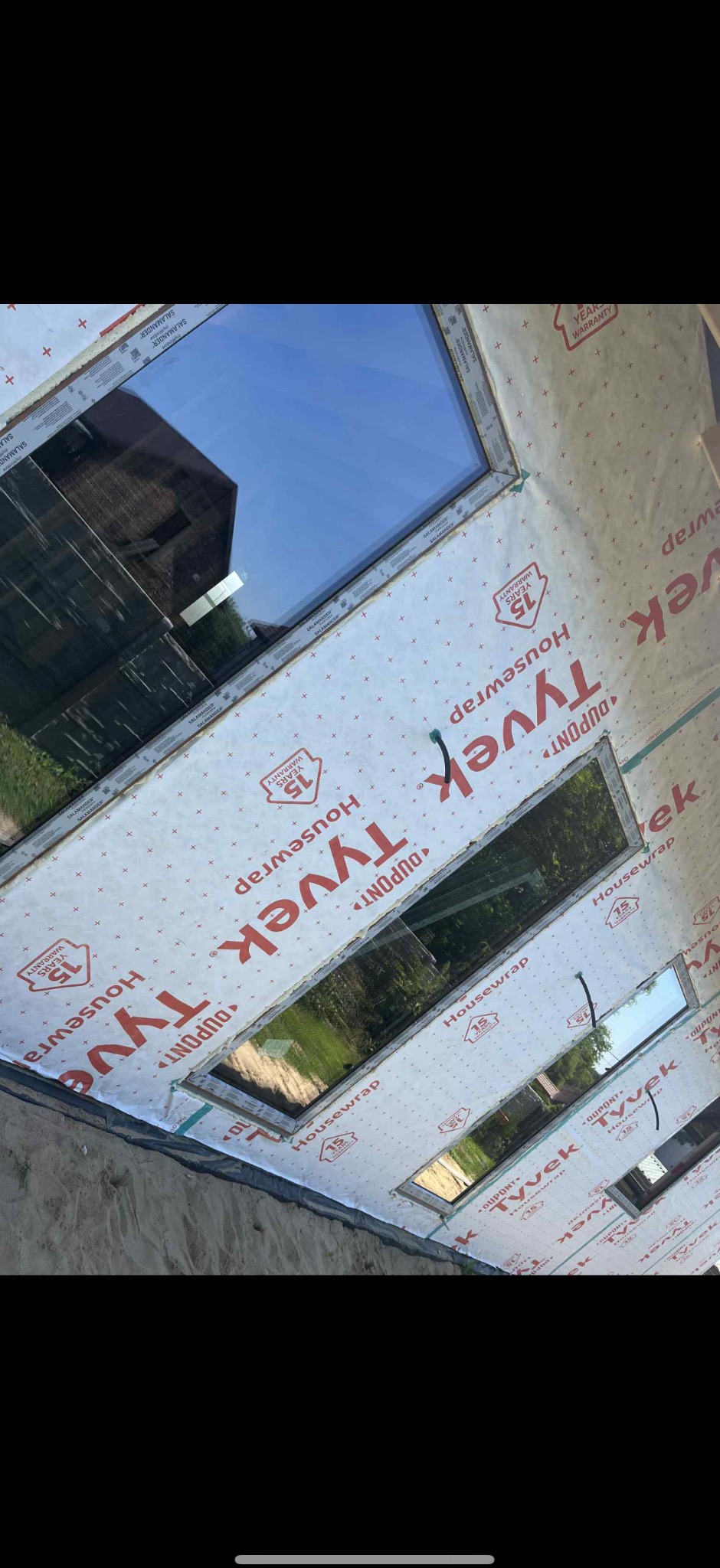 Montaż okien dachowych w warstwie hydroizolacji Tyvek Housewrap. Widoczne ramy okienne Salamander i odbicie krajobrazu w szybach. Prace budowlane w toku.