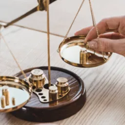 Złote wagi szalkowe z odważnikami; dłoń dodaje ciężarek. Symbol sprawiedliwości, równowagi i uczciwości w kontekście finansów i kredytów.