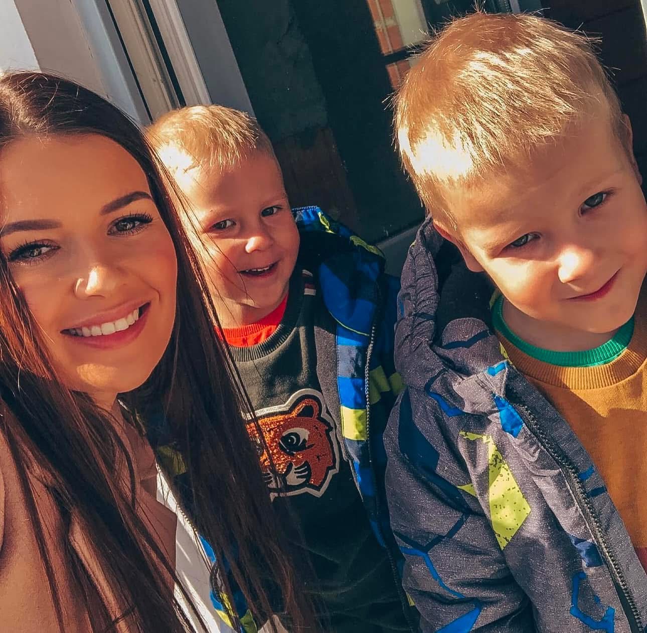 Uśmiechnięta kobieta robi selfie z dwójką roześmianych chłopców w kurtkach na tle jasnego okna. Widoczne detale ubrań i promienny wyraz twarzy.