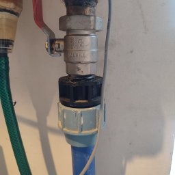 Yury Shauliukevich Njuthor Hydraulik - Szczegółowe ujęcie zaworu kulowego z czerwoną rączką, połączonego z niebieską rurą plastikową i zielonym wężem, na tle jasnej ściany z widocznymi cieniami.