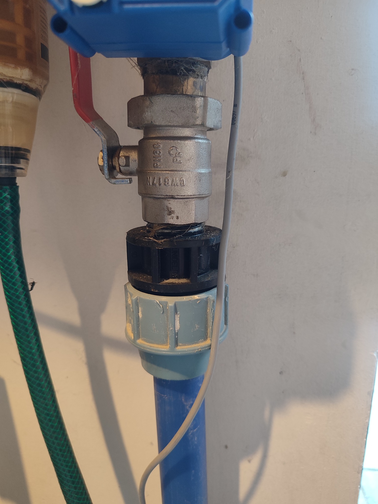 Szczegółowe ujęcie zaworu kulowego z czerwoną rączką, połączonego z niebieską rurą plastikową i zielonym wężem, na tle jasnej ściany z widocznymi cieniami.