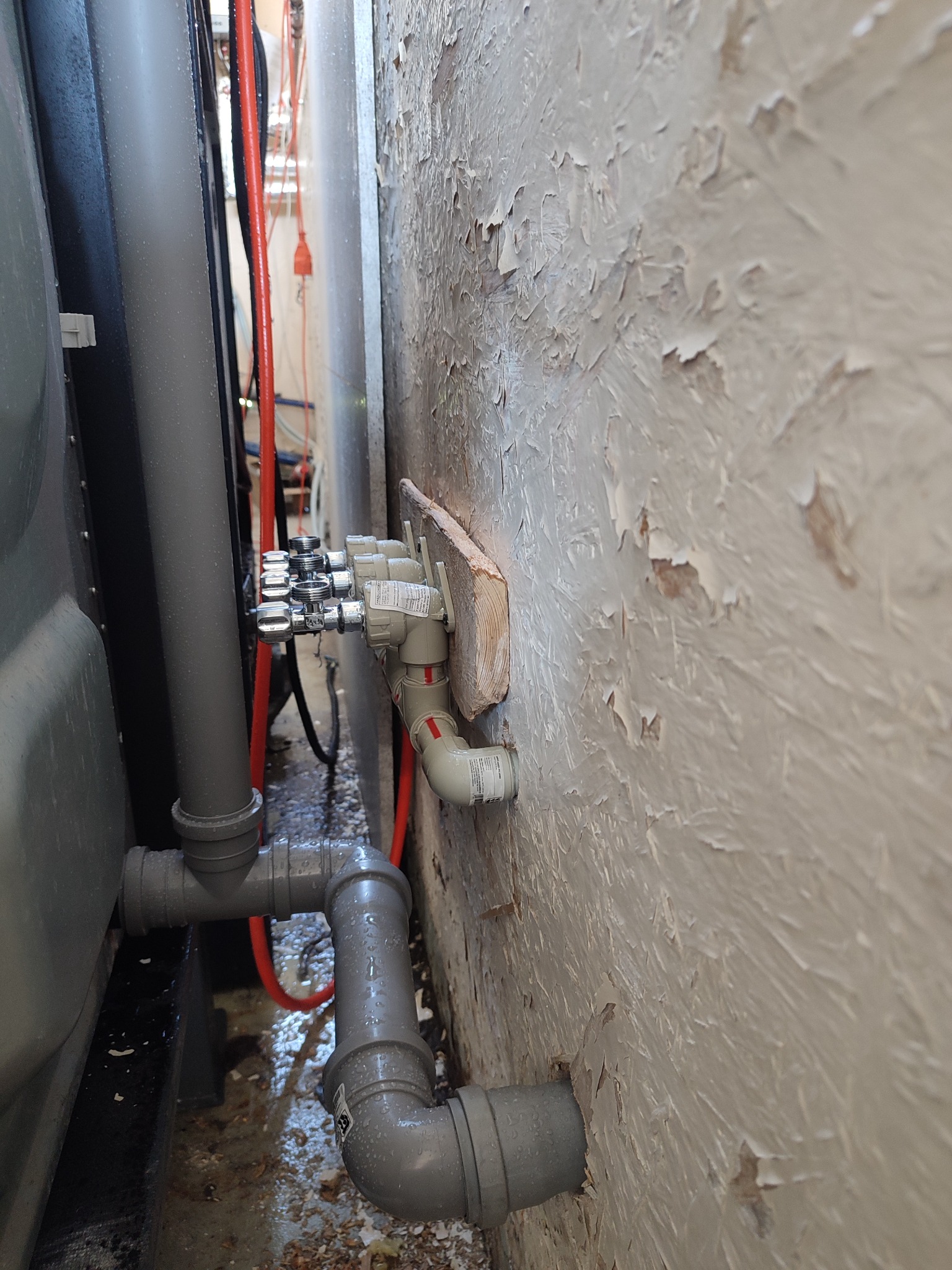 Szare rury i zawory instalacji wodnej zamontowane przy ścianie z łuszczącą się farbą. Widoczne złącza i elementy instalacji, z bliska, w ciasnej przestrzeni.