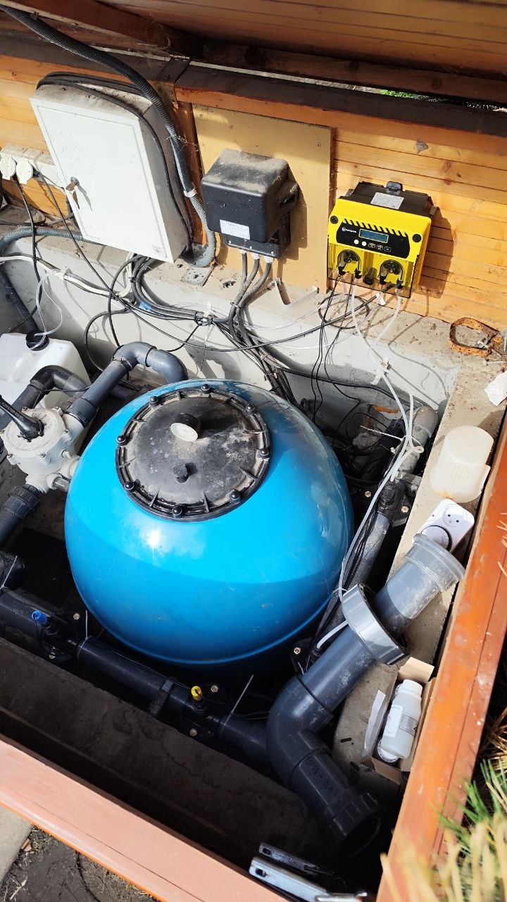 Niebieski filtr piaskowy do basenu z czarną pokrywą, otoczony instalacją hydrauliczną i przewodami elektrycznymi w drewnianej obudowie.