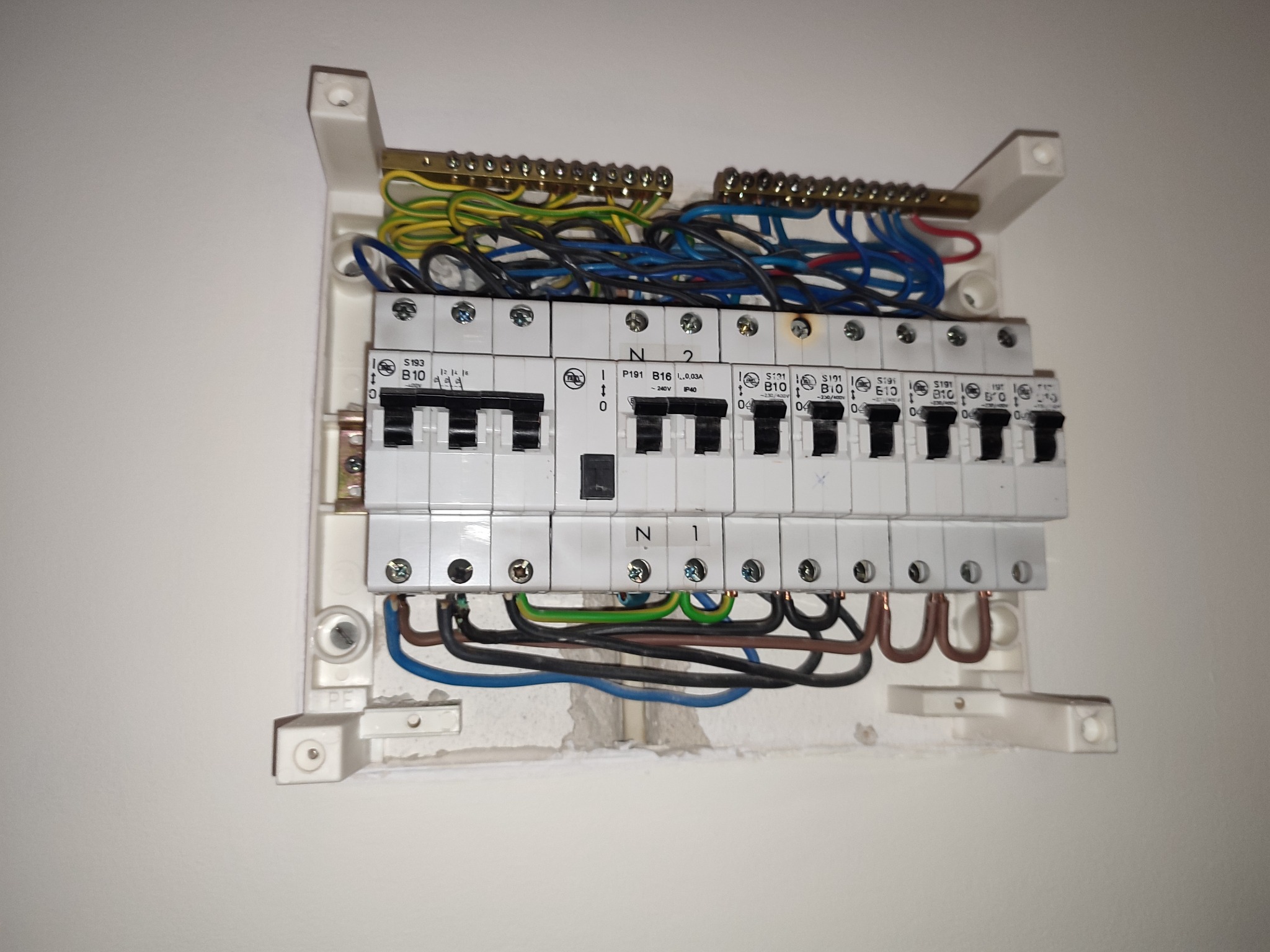 Rozdzielnica elektryczna z bezpiecznikami i okablowaniem, zamontowana na ścianie. Widoczne przewody w różnych kolorach i elementy mocujące.