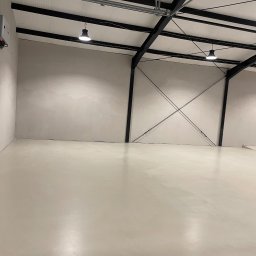 Karpowicz-Korbwaren - Jasna, gładka posadzka w hali przemysłowej z metalową konstrukcją stropu i oświetleniem. Widoczna brama garażowa z lewej strony. Świeżo wykonana wylewka.