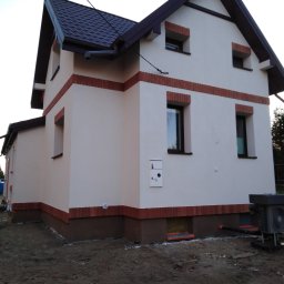 Radbud - Ocieplony dom jednorodzinny z elewacją w kolorze kremowym, z brązowymi akcentami ceglanymi i ciemnym dachem. Widoczny fragment gruntu przed budynkiem.