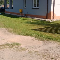 Radbud - Dom jednorodzinny z szarą elewacją, brązowym dachem i ceglaną podmurówką. Widoczne dwa okna z żaluzjami i żółta skrzynka gazowa. Trawa i piaszczysta droga.