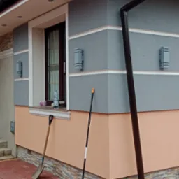 Wykończona elewacja domu z szarą i brzoskwiniową kolorystyką, widoczne oświetlenie zewnętrzne, rynna i narzędzia ogrodowe opierające się o ścianę.