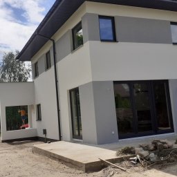 Radbud - Dwukondygnacyjny dom po odnowieniu elewacji w odcieniach szarości i bieli. Na rusztowaniu malarz w pomarańczowej koszulce. Widoczne materiały budowlane.