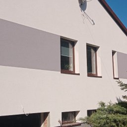 Radbud - Dwukolorowa elewacja domu z szarą strefą. Widoczne okna z brązowymi ramami, rusztowanie i fragment osoby. Tło stanowi błękitne niebo i zieleń drzew.