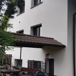 Biała elewacja domu po malowaniu, z brązowymi elementami wykończeniowymi, widok z boku na tle zieleni i narzędzi malarskich. Świeżo pomalowana elewacja.