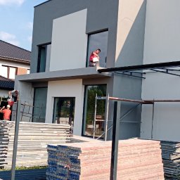 Radbud - Dwóch pracowników maluje dwukolorową elewację domu z rusztowania. Na pierwszym planie stosy materiałów budowlanych i elementy konstrukcyjne.