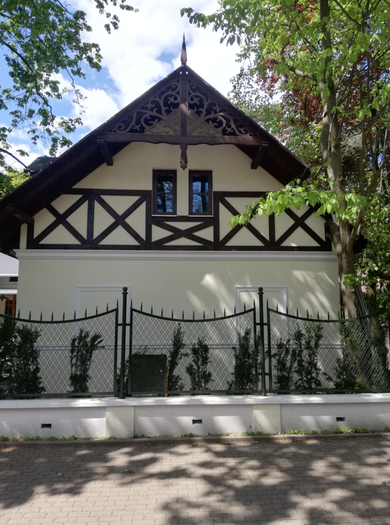 Elewacja domu jednorodzinnego z dekoracyjnymi elementami drewnianymi w Drzewicy. Jasna fasada, ciemne okna i brama, ozdobne wykończenie dachu.