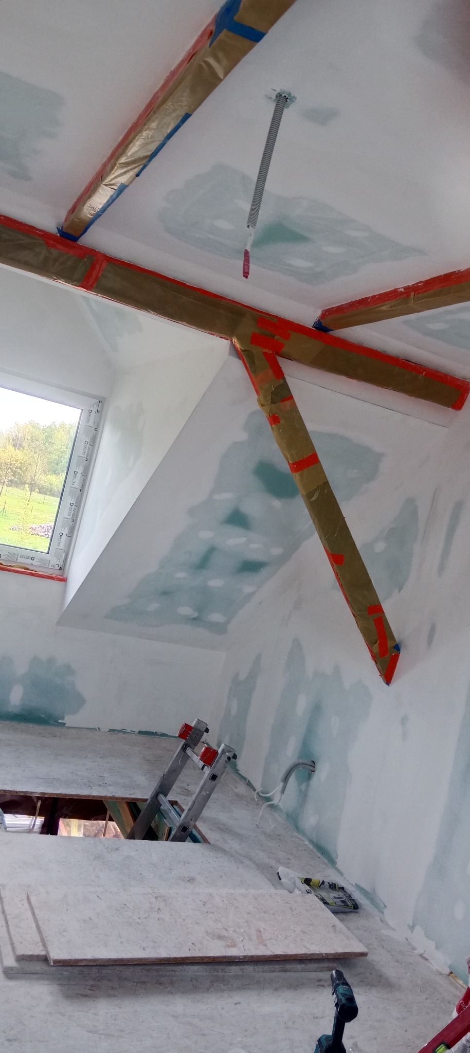 Skośne poddasze w trakcie prac wykończeniowych: ściany i sufit pokryte gładzią gipsową, drewniane elementy zabezpieczone folią, drabina i narzędzia na podłodze.