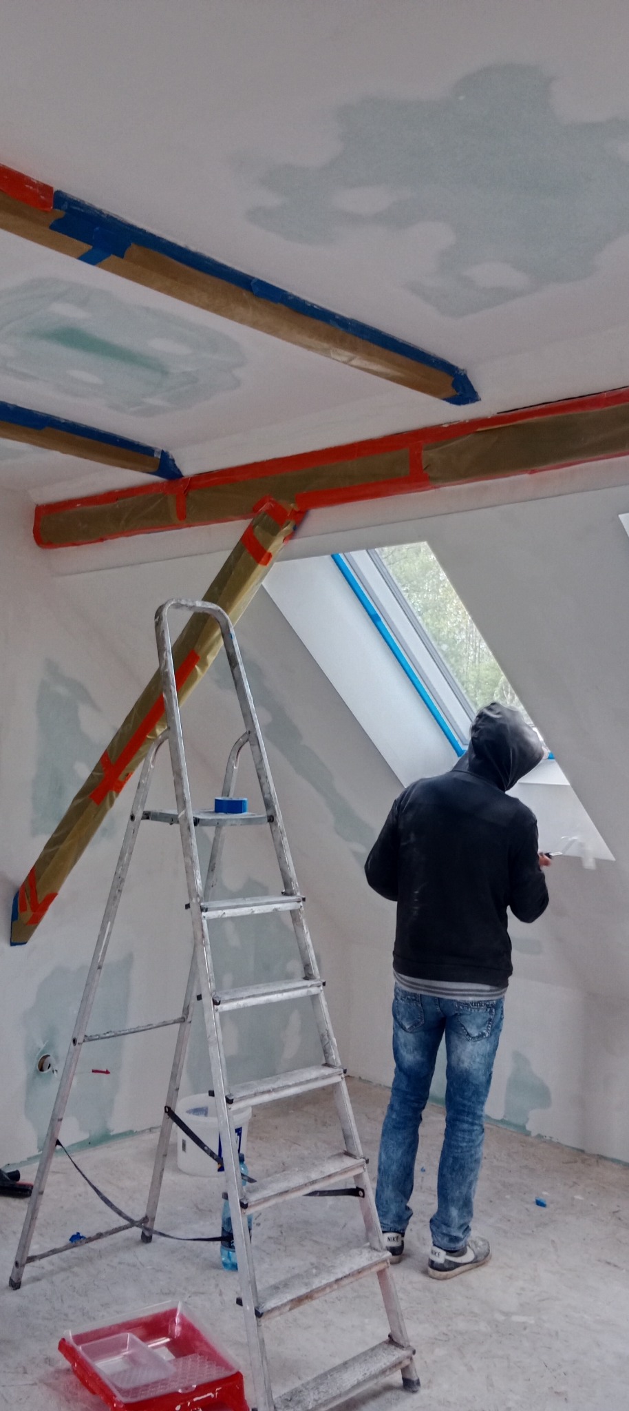 Prace malarskie na poddaszu: mężczyzna maluje ścianę przy oknie dachowym, drabina oparta o belkę zabezpieczoną folią i taśmą. Widoczne narzędzia malarskie.