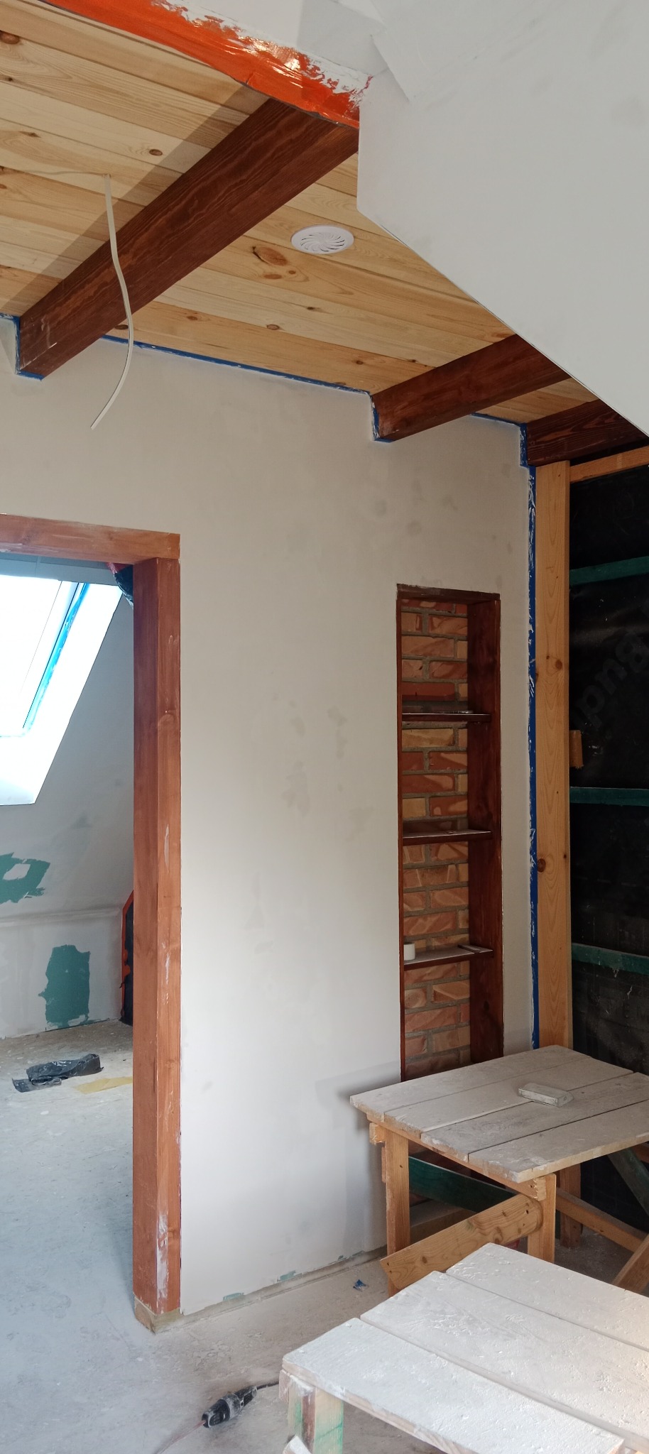 Wnętrze w trakcie prac wykończeniowych: otynkowana ściana z wnęką, drewniany sufit z belkami, stolarka drzwiowa i okienna, widoczne narzędzia i rusztowania malarskie.