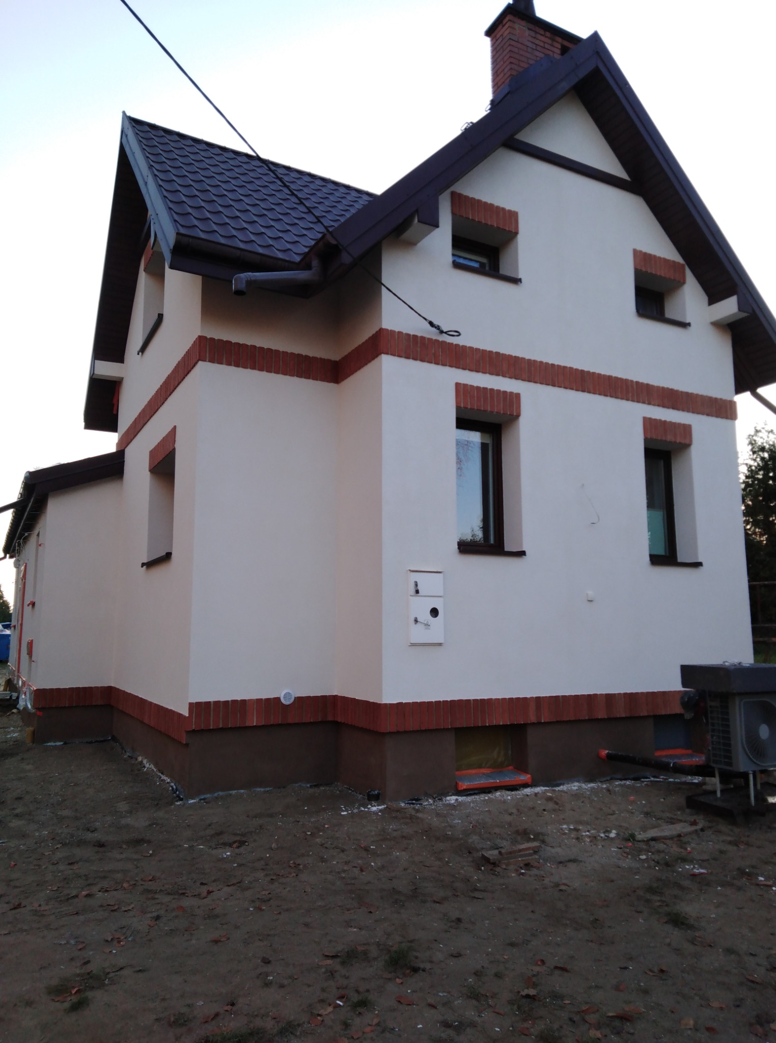 Ocieplony dom jednorodzinny z elewacją w kolorze kremowym, z brązowymi akcentami ceglanymi i ciemnym dachem. Widoczny fragment gruntu przed budynkiem.