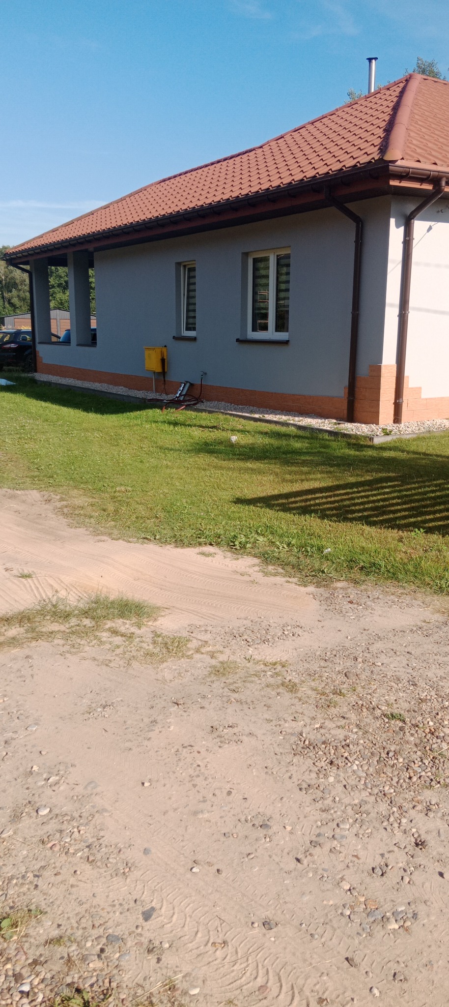 Dom jednorodzinny z szarą elewacją, brązowym dachem i ceglaną podmurówką. Widoczne dwa okna z żaluzjami i żółta skrzynka gazowa. Trawa i piaszczysta droga.