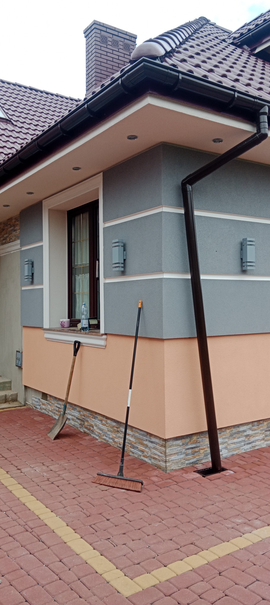 Wykończona elewacja domu z szarą i brzoskwiniową kolorystyką, widoczne oświetlenie zewnętrzne, rynna i narzędzia ogrodowe opierające się o ścianę.