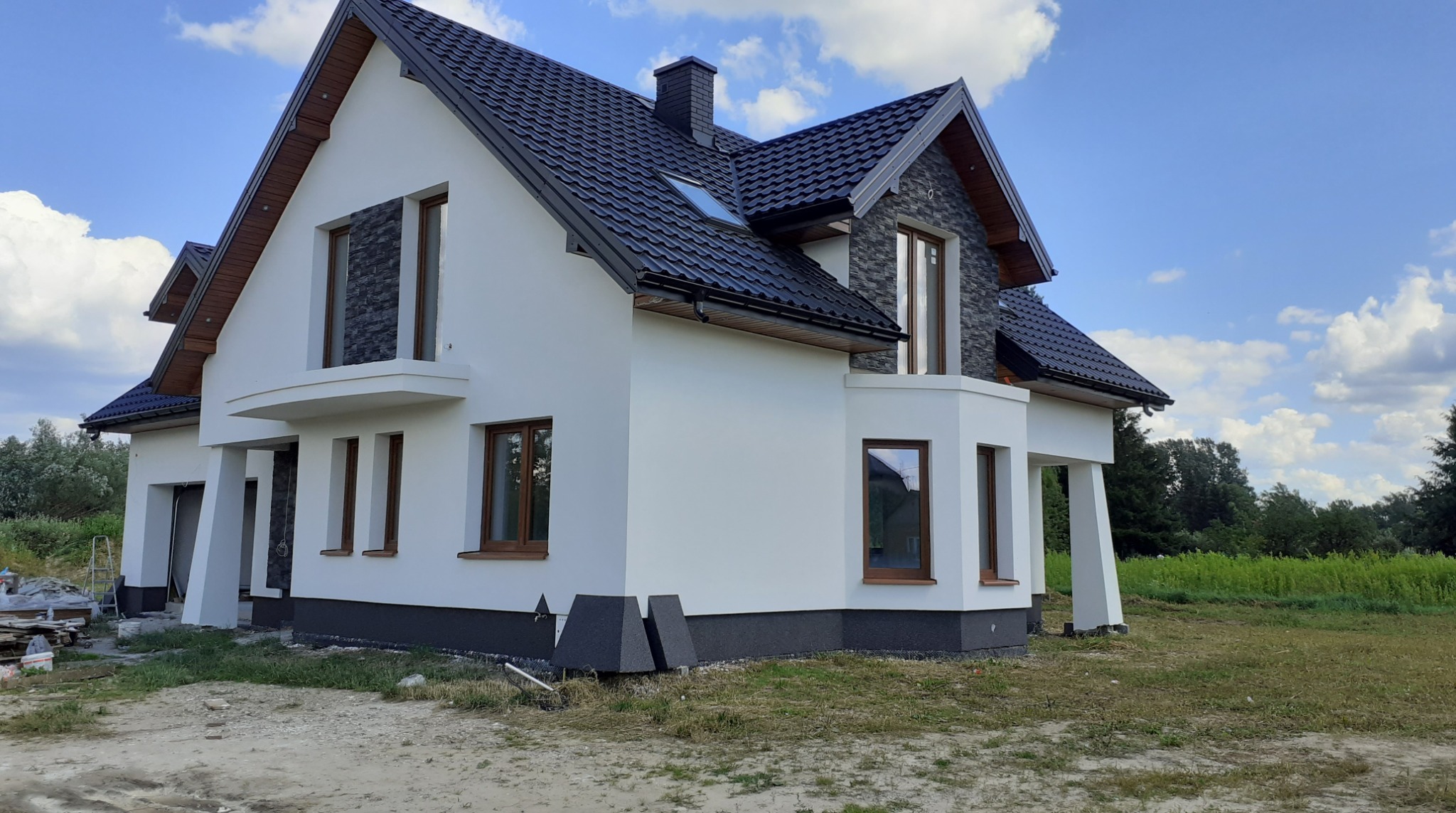 Nowoczesny dom jednorodzinny z białą elewacją, czarną dachówką i elementami kamiennymi. Widok z boku, w tle błękitne niebo z chmurami i zielone drzewa.