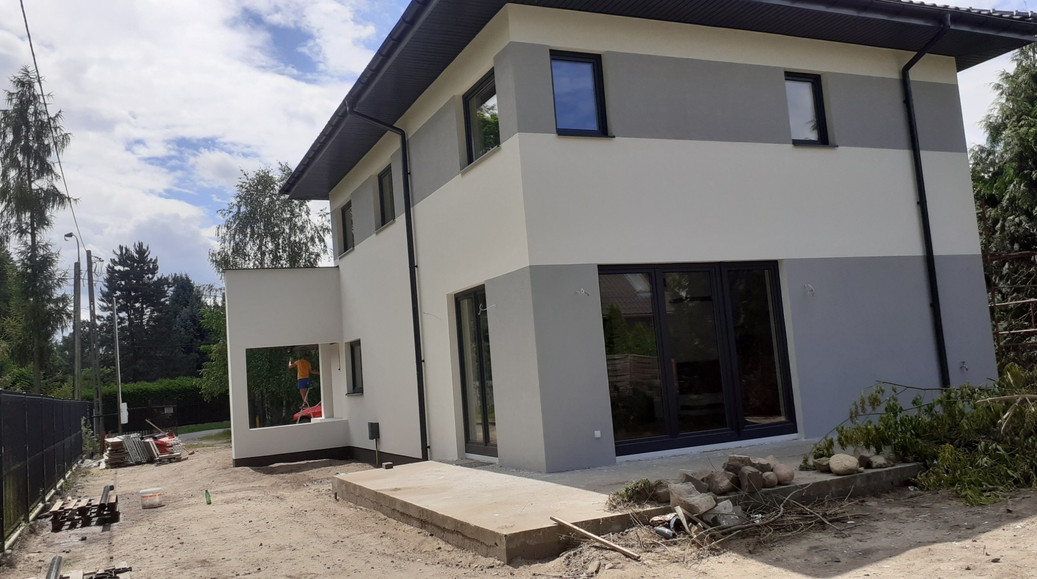 Dwukondygnacyjny dom po odnowieniu elewacji w odcieniach szarości i bieli. Na rusztowaniu malarz w pomarańczowej koszulce. Widoczne materiały budowlane.