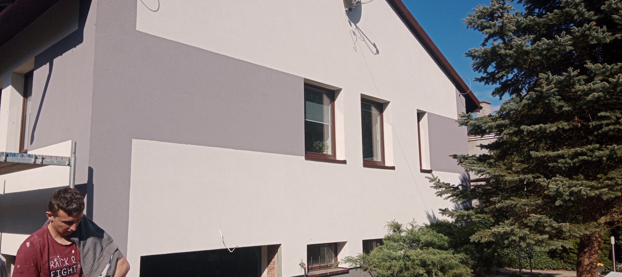 Dwukolorowa elewacja domu z szarą strefą. Widoczne okna z brązowymi ramami, rusztowanie i fragment osoby. Tło stanowi błękitne niebo i zieleń drzew.