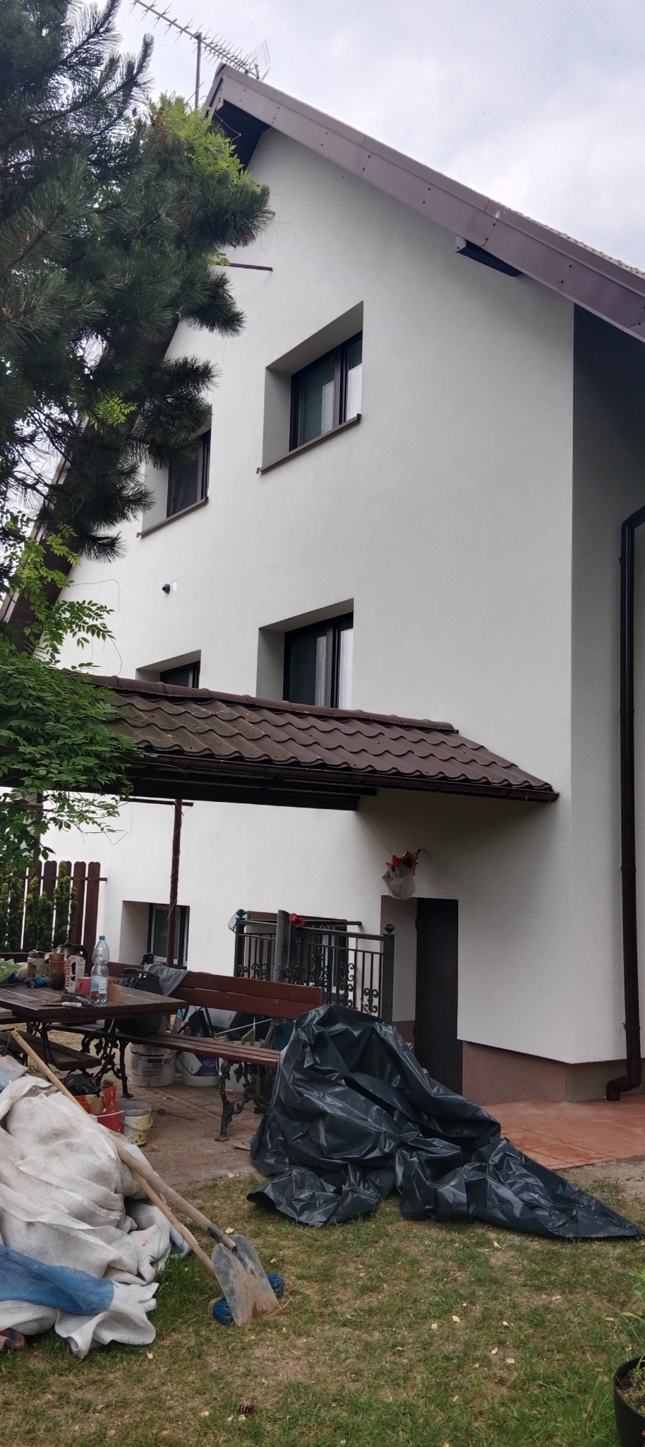 Biała elewacja domu po malowaniu, z brązowymi elementami wykończeniowymi, widok z boku na tle zieleni i narzędzi malarskich. Świeżo pomalowana elewacja.