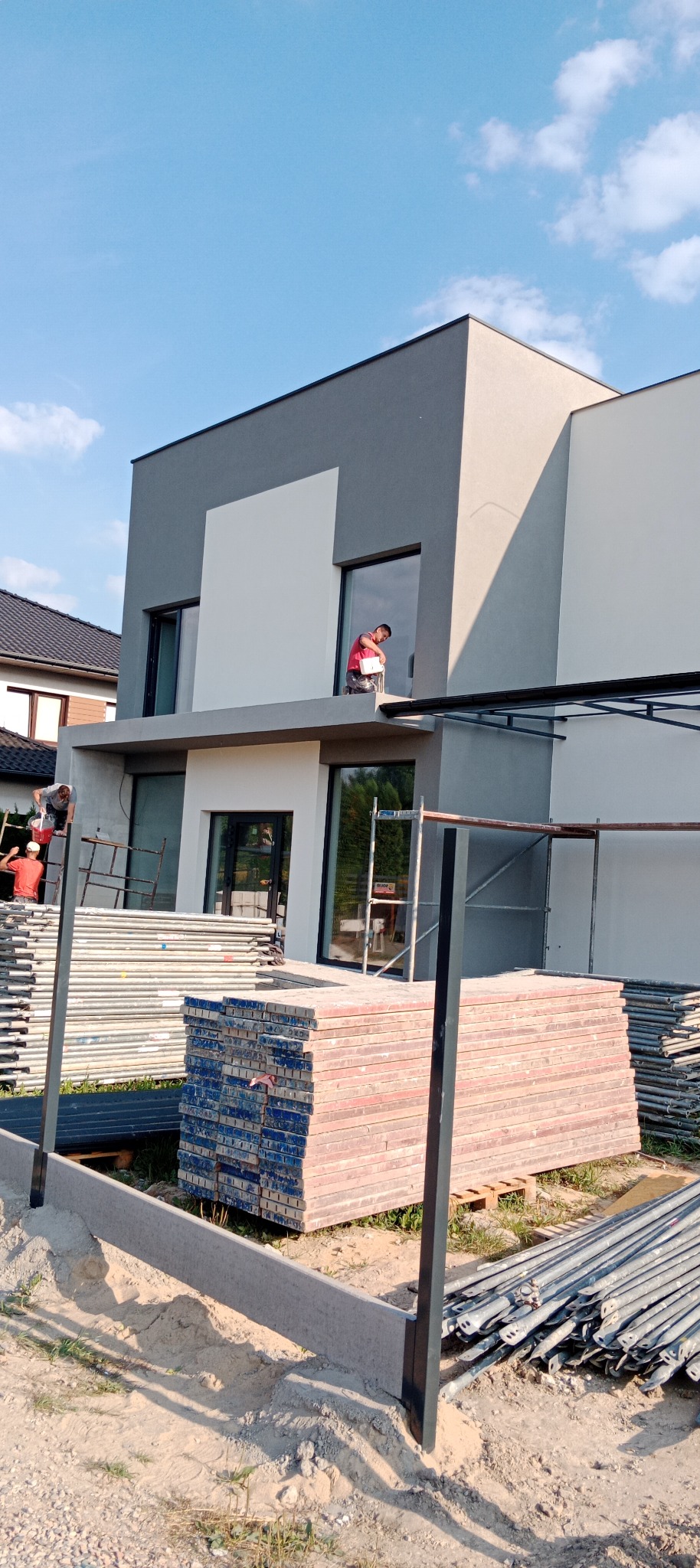 Dwóch pracowników maluje dwukolorową elewację domu z rusztowania. Na pierwszym planie stosy materiałów budowlanych i elementy konstrukcyjne.