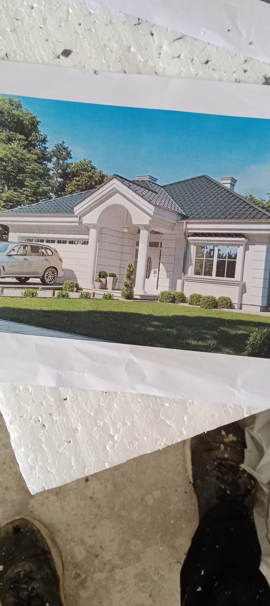 Wydruk wizualizacji domu jednorodzinnego z garażem i samochodem, leżący na styropianie. Widoczny fragment buta. Nietypowe ujęcie.