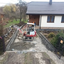 Geo grunt serwis Kraków - Mini koparka na placu budowy, przygotowuje podłoże z gruzu i kamieni pod chodnik lub podjazd. Widoczne obrzeża i dom w tle. Prace ziemne w toku.