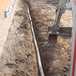 Geo grunt serwis Kraków - Wkopana rura w ziemi obok budynku, widoczna koparka. Etap prac ziemnych z użyciem ciężkiego sprzętu. Na pierwszym planie fragment koparki i wykopana ziemia.