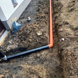 Geo grunt serwis Kraków - Wykop z ułożonymi rurami (pomarańczowa i czarna) przy fundamencie budynku. Widoczny fragment elewacji i okna. Grunt ziemny, przygotowany pod dalsze prace.