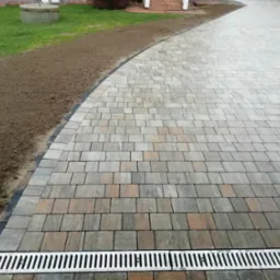 Ułożona kostka brukowa w odcieniach szarości i brązu, z widocznym odpływem liniowym i przygotowanym terenem pod trawnik. Nowoczesne i estetyczne wykończenie podjazdu.