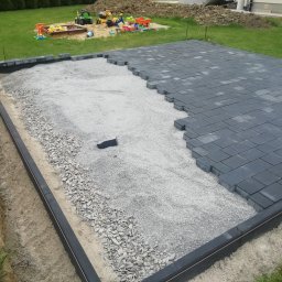 Geo grunt serwis Kraków - Częściowo ułożona, szara kostka brukowa na podsypce z kruszywa. Widoczne obrzeża i przygotowane podłoże. W tle trawnik i zabawki dziecięce.