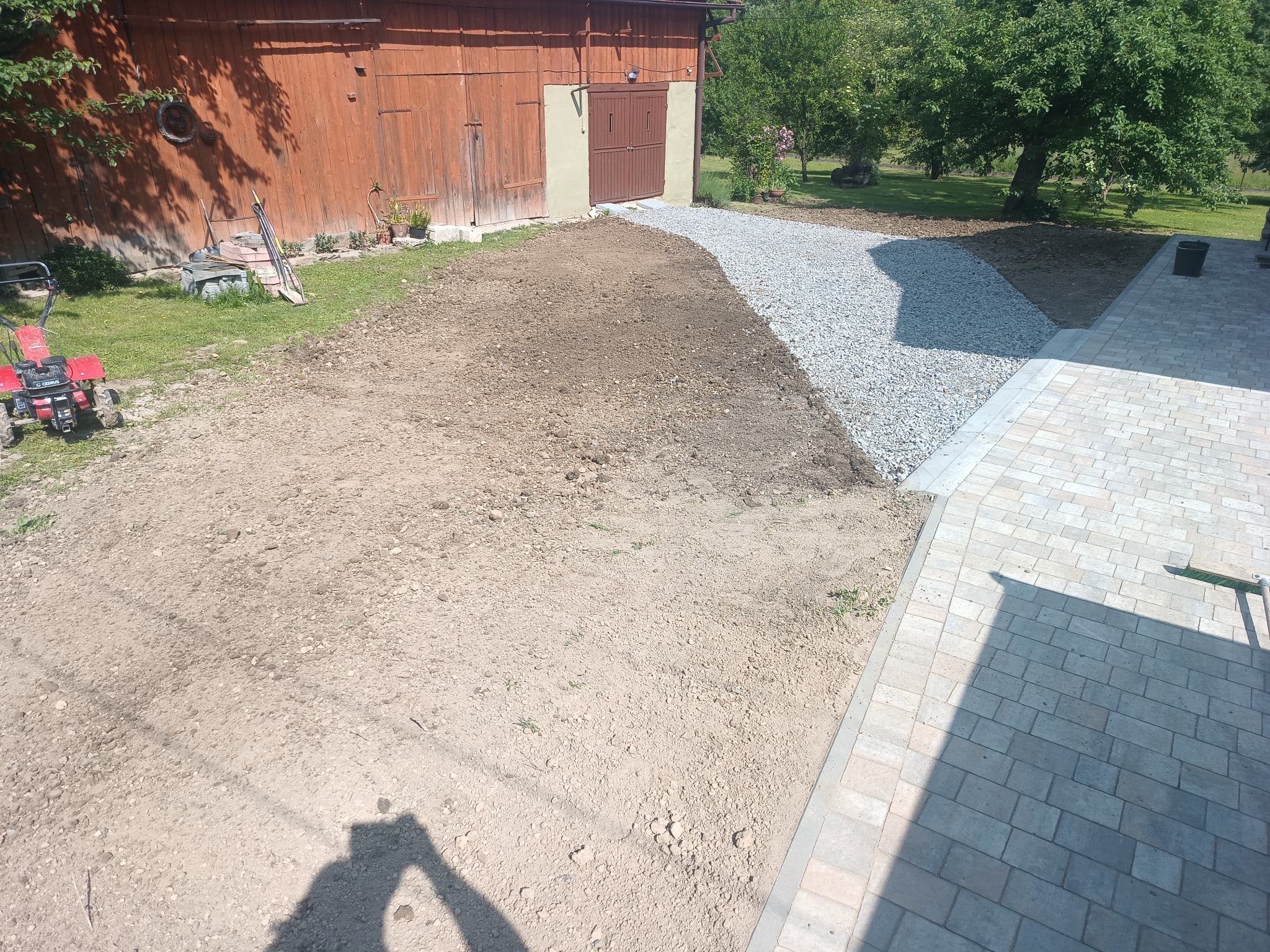 Przygotowany teren pod trawnik z fragmentem ścieżki z kamienia i kostki brukowej. W tle budynek gospodarczy z drewnianymi drzwiami. Widoczne narzędzia i maszyna.