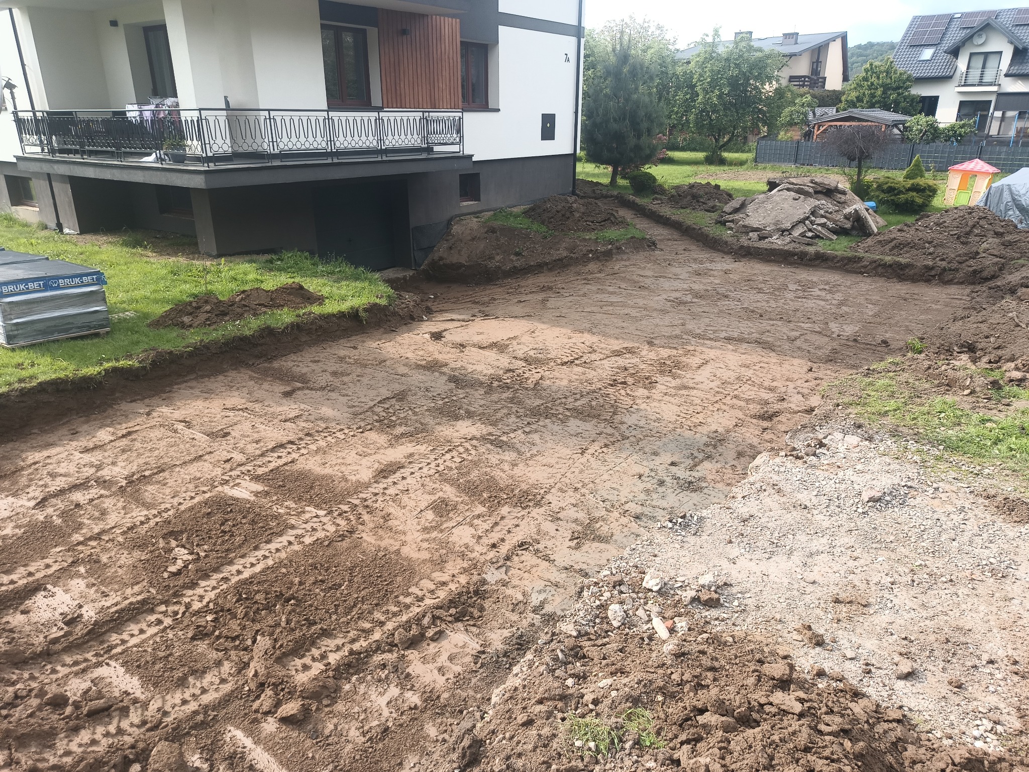 Przygotowany teren pod kostkę brukową przed nowoczesnym domem. Widoczne ślady opon i paleta z kostką. W tle ogród i sąsiednie budynki.