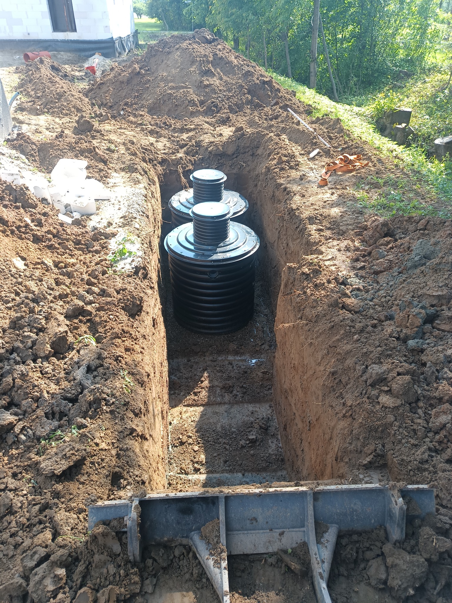 Wykop pod szambo z trzema zbiornikami z tworzywa sztucznego w ziemi. Widoczny fragment budynku w tle i rów wykopany koparką. Słoneczny dzień.